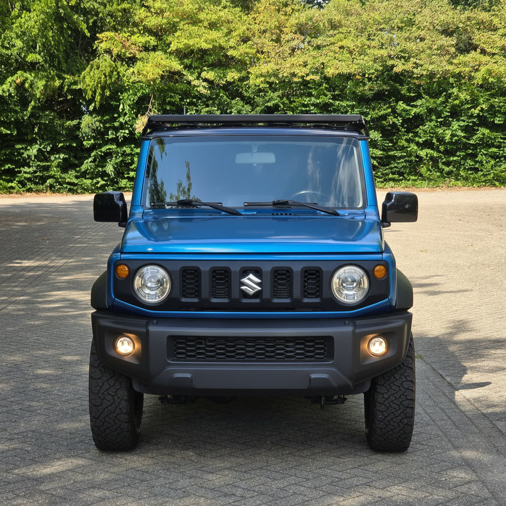 A blue Suzuki Jimny off-road vehicle with a black wildpeak roof rack and front bumper, parked on a paved surface with a green forest background.