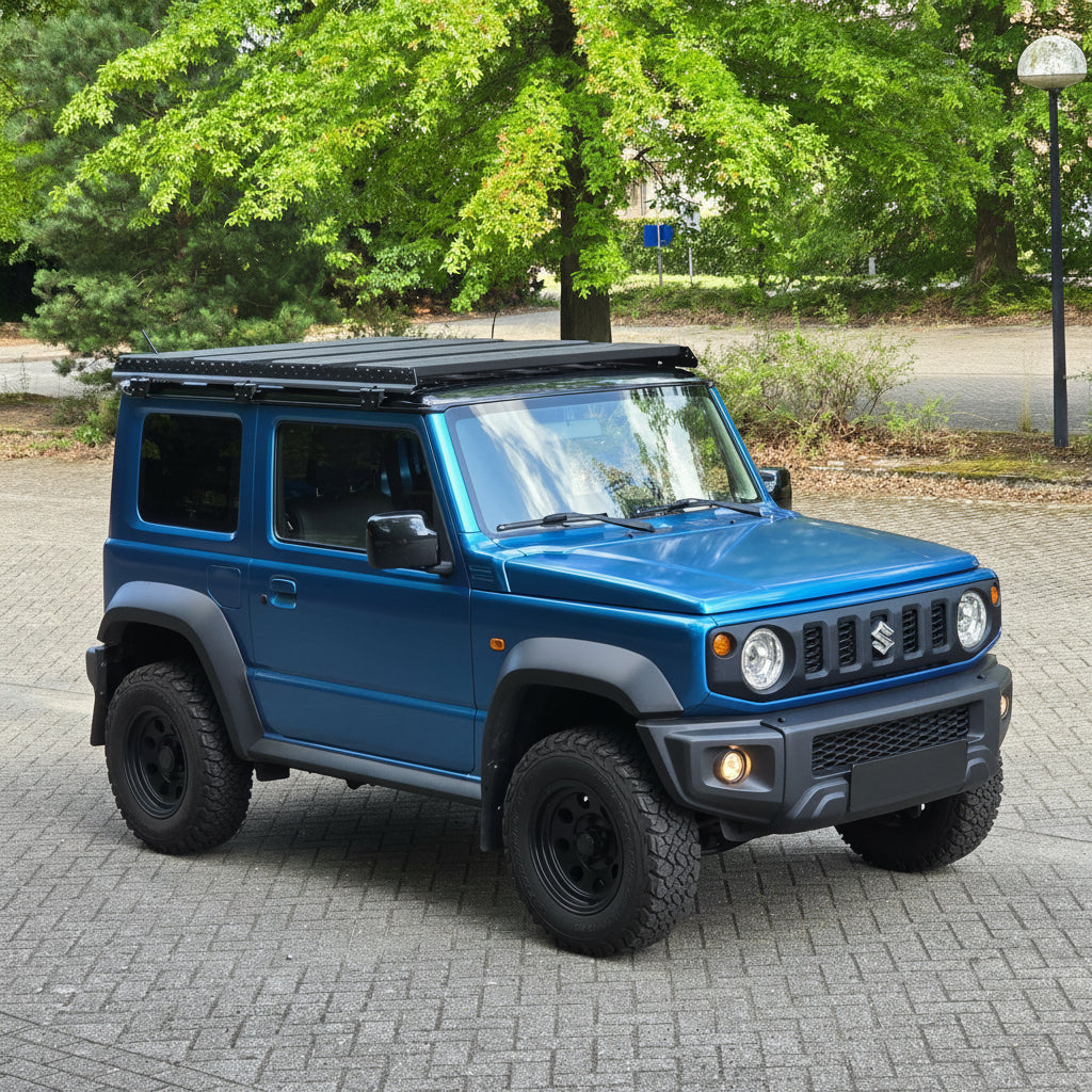 A blue Suzuki Jimny SUV with a black wildpeak roof rack and off-road tires, parked on a paved street with trees in the background.